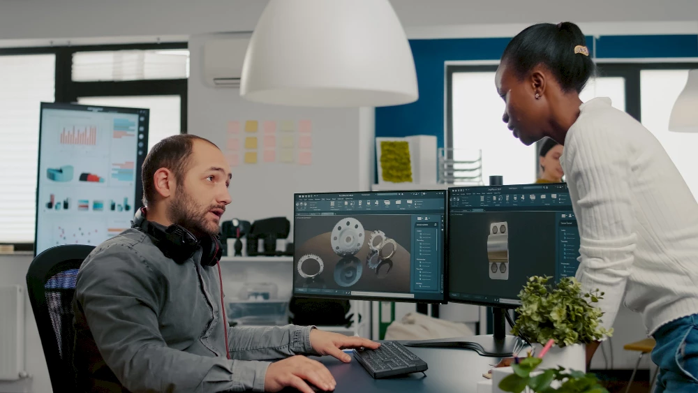 Conversations between employees at a desk. CAD models can be seen on screens in the background.